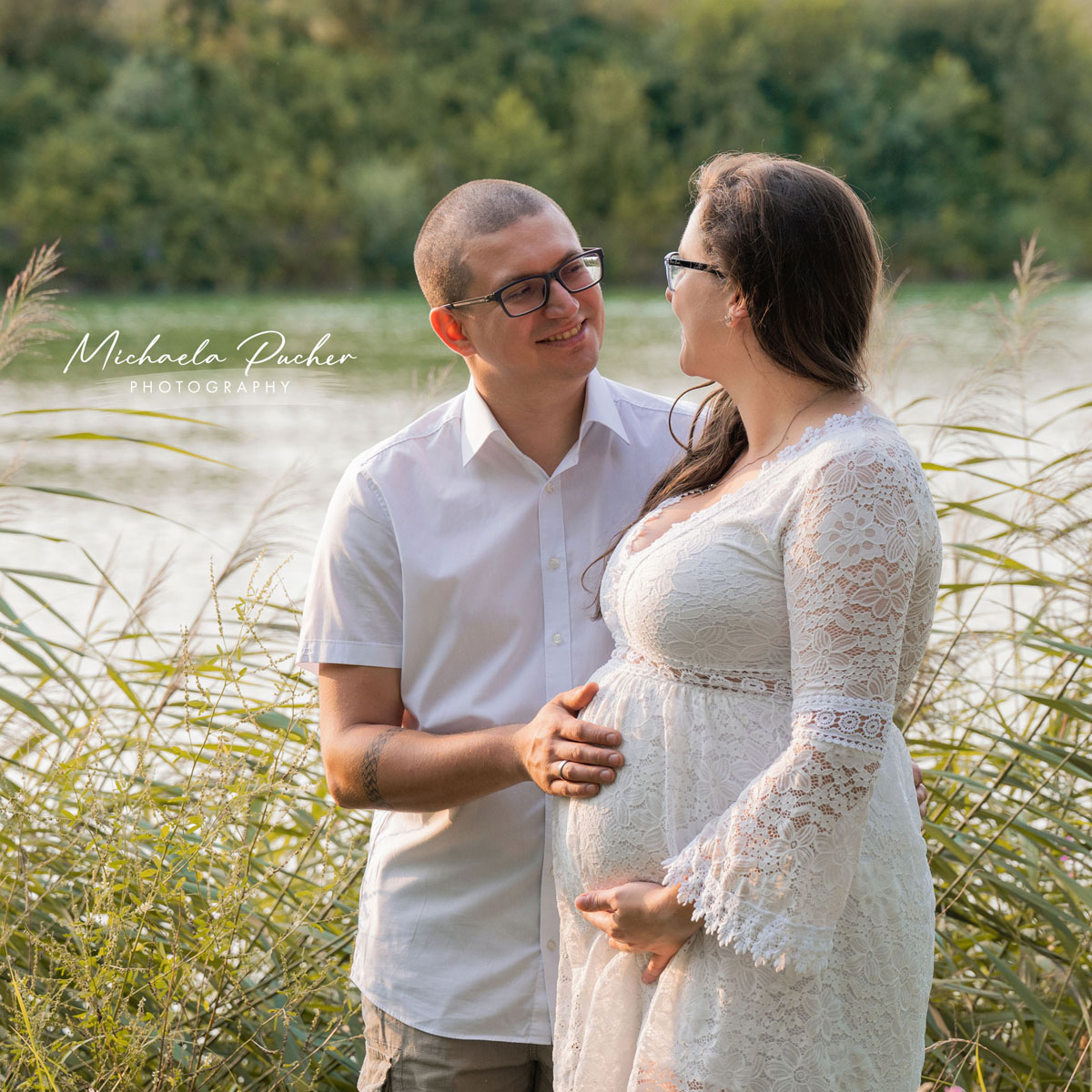 Paar steht am Seeufer im warmen Abendlicht, der Mann hält liebevoll den Babybauch seiner Partnerin, beide lächeln sich innig an.